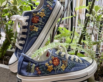 Può includere: Un paio di sneakers in tela blu con suole bianche e ricamo floreale colorato. Il ricamo presenta fiori, foglie e alberi in tonalità di rosso, arancione, giallo e verde.