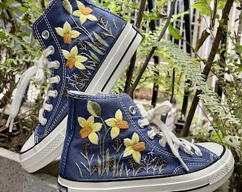 May include: A pair of blue high-top canvas sneakers with white soles and laces. The sneakers are embroidered with yellow daffodils and green leaves.