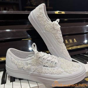 May include: White canvas sneakers with intricate floral beadwork and ribbon laces. The shoes feature a low-top design with a white rubber sole, perfect for a wedding or special occasion. The Vans logo is visible on the side.