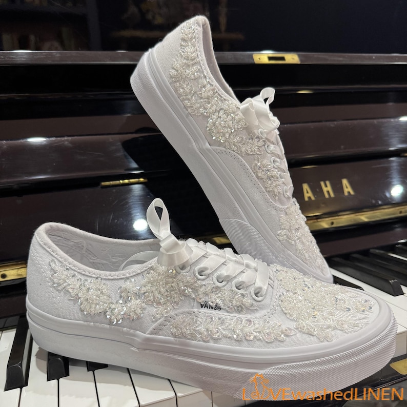 May include: White canvas sneakers with intricate floral beadwork and ribbon laces. The shoes feature a low-top design with a white rubber sole, perfect for a wedding or special occasion. The Vans logo is visible on the side.