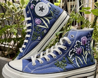 May include: A pair of blue high-top Converse sneakers with white laces and a white rubber sole. The sneakers are decorated with embroidered flowers and leaves in shades of green, purple, and white.