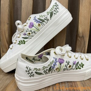 Può includere: Sneakers bianche con piattaforma e ricami floreali, con il testo "Mrs. Dunton" sul lato.