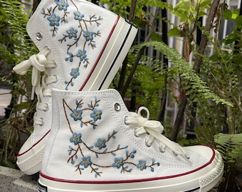 May include: White high-top canvas sneakers with blue floral embroidery. The shoes have a white rubber sole and a red trim around the top.