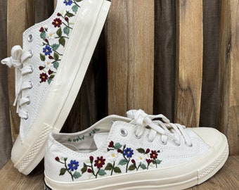 Op de afbeelding: Witte canvas lage sneakers met geborduurde bloemen in rood, blauw, groen en wit. De schoenen hebben witte veters en een witte rubberen zool.