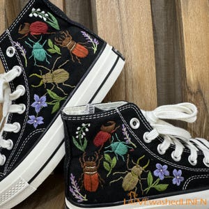 Könnte beinhalten: Schwarze Canvas-High-Top-Sneaker mit bunten gestickten Insekten und Blumen. Die Schuhe haben weiße Schnürsenkel und eine weiße Gummisohle.