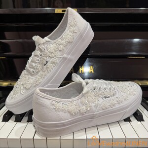 May include: White Vans sneakers embellished with floral lace and beadwork. The shoes have white laces and a white sole. The Vans logo is visible on the side of the shoe. The shoes are displayed on a piano keyboard.