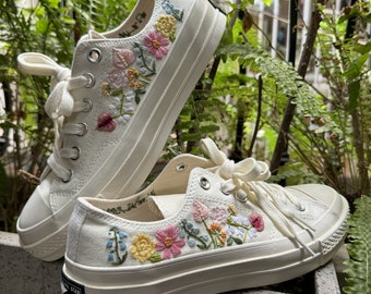 May include: A pair of white canvas sneakers with floral embroidery. The embroidery features pink, yellow, blue, and green flowers. The sneakers have a white rubber sole and a black star logo on the side.