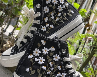 May include: A pair of black high-top canvas sneakers with white daisy embroidery. The sneakers have white laces and a white rubber sole.