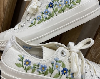 May include: Cream-colored canvas sneakers with embroidered blue and yellow flowers and green leaves. The low-top shoes have white laces and a thick, off-white rubber sole. The floral embroidery adds a decorative touch to the casual footwear.