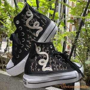 May include: Black high-top sneakers with white embroidered snakes and branches on the sides. The sneakers have white soles and black laces.