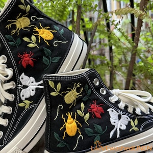 Könnte beinhalten: Schwarze High-Top-Canvas-Schuhe mit gestickten Insekten und Blumen. Die Schuhe haben weiße Schnürsenkel und eine weiße Gummisohle.