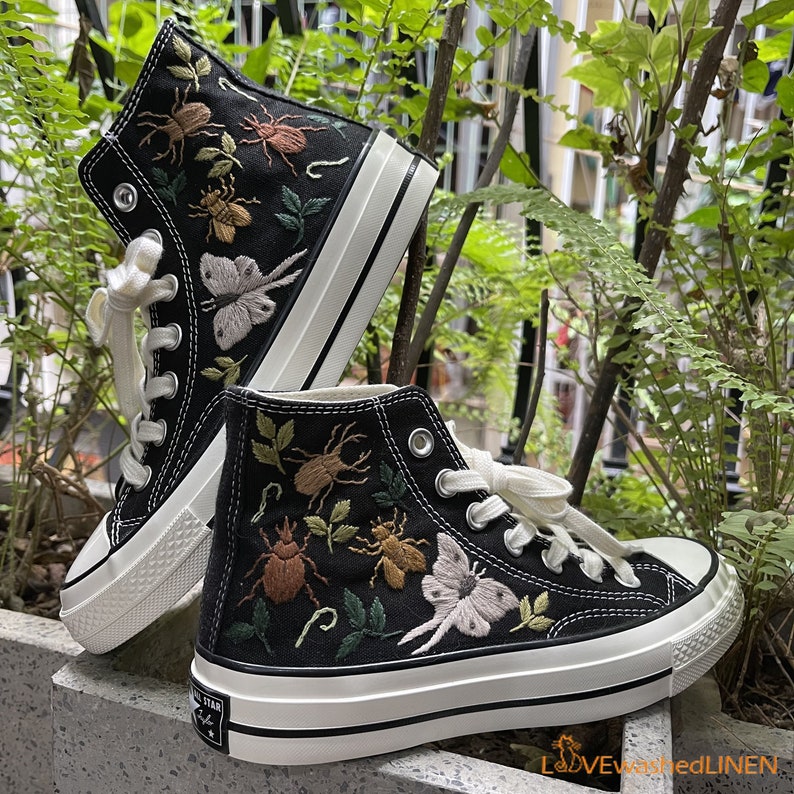 May include: Black high-top canvas sneakers with white soles and embroidered insects and flowers. The sneakers have a vintage look.