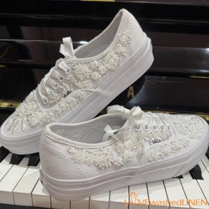 May include: White canvas sneakers with floral embellishments and satin ribbon laces. The shoes feature a thick white sole and are decorated with beaded floral designs. The Vans logo is visible on the side of the shoe.