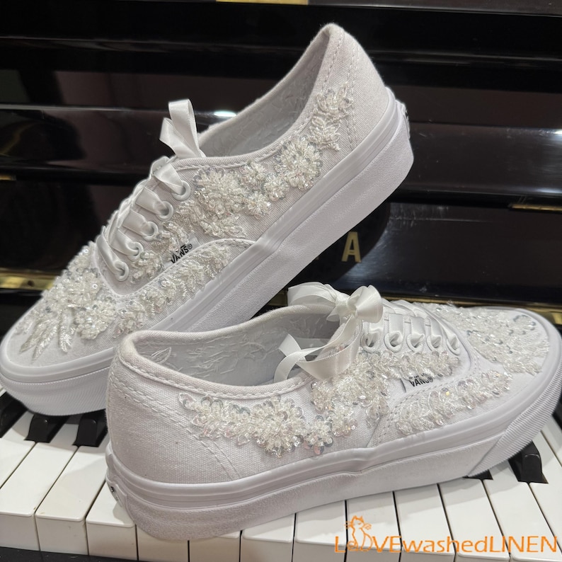 May include: White canvas sneakers with floral embellishments and satin ribbon laces. The shoes feature a thick white sole and are decorated with beaded floral designs. The Vans logo is visible on the side of the shoe.