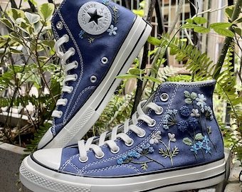 May include: A pair of blue high-top Converse sneakers with white soles and laces. The sneakers are decorated with embroidered flowers in shades of blue and white.
