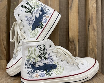 Puede incluir: Zapatillas blancas de lona de caña alta con tiburones azules y algas verdes bordadas. Las zapatillas tienen una suela de goma blanca y costuras rojas.