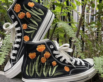 May include: Black high-top sneakers with embroidered orange flowers and green leaves. The sneakers have white laces, a white rubber sole, and a black stripe. The text "LOVEwashedLINEN" is visible on the side.