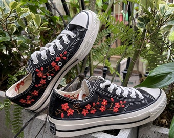 May include: Black canvas sneakers with white laces and red embroidered flowers. The shoes have a white rubber sole.