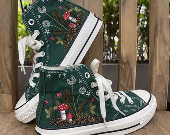 Puede incluir: Un par de zapatillas altas de lona verde con cordones blancos y suela de goma blanca. Las zapatillas están bordadas con un diseño floral que incluye setas rojas, hojas verdes y dientes de león blancos.