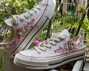May include: A pair of white canvas sneakers with red, purple, and green embroidered flowers and leaves. The sneakers have a white rubber sole and a red stripe around the top.