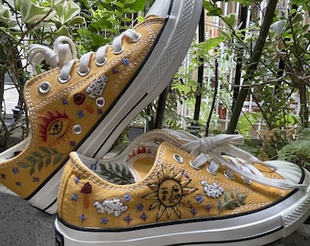 Può includere: Un paio di sneakers in tela gialla con suole bianche, dipinte a mano con dettagli ricamati di un sole, nuvole e fiori.