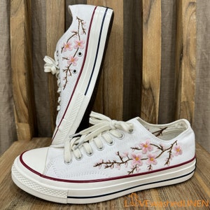 Könnte beinhalten: Weiße Canvas-Sneaker mit rosa gestickten Kirschblüten an der Seite. Die Schuhe haben eine weiße Gummisohle und einen roten Streifen um die Oberseite.