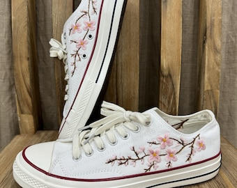 May include: White canvas sneakers with pink embroidered cherry blossoms on the side. The shoes have a white rubber sole and a red stripe around the top.