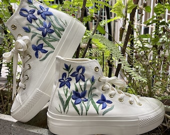 May include: White high-top canvas sneakers with blue and green embroidered flowers. The sneakers have a thick white platform sole.