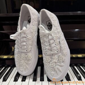 May include: A pair of white Vans sneakers, heavily embellished with floral embroidery and beadwork. The shoes feature white laces tied in bows and are set against a piano keyboard. The Vans logo is visible on the side.
