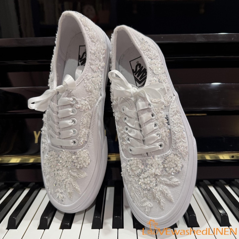 May include: A pair of white Vans sneakers, heavily embellished with floral embroidery and beadwork. The shoes feature white laces tied in bows and are set against a piano keyboard. The Vans logo is visible on the side.
