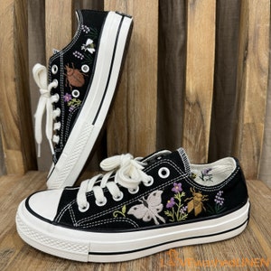 Könnte beinhalten: Schwarze Canvas-Sneaker mit weißen Schnürsenkeln und einer weißen Sohle. Die Schuhe sind mit einem Schmetterling, Blumen und einem Käfer bestickt.