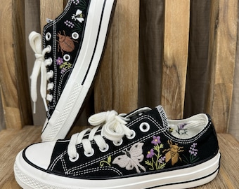 May include: Black canvas sneakers with white laces and a white sole. The shoes are embroidered with a butterfly, flowers, and a beetle.