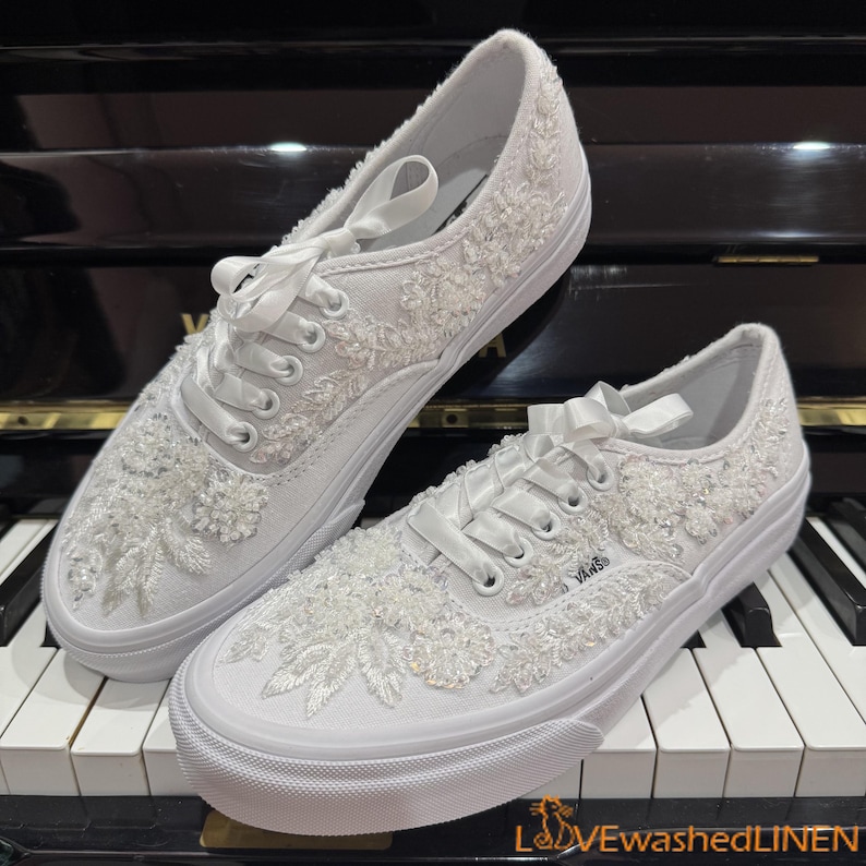 May include: A pair of white Vans sneakers, embellished with intricate floral lace and beadwork. The shoes feature white laces and a white rubber sole. The design includes a satin ribbon bow at the top of the laces. The Vans logo is visible on the side.