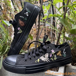 Könnte beinhalten: Ein Paar schwarze Canvas-Sneaker mit gestickten Blumendesigns, darunter Schmetterlinge, Käfer und Blumen. Die Sneaker haben weiße Schnürsenkel und eine schwarze Gummisohle.