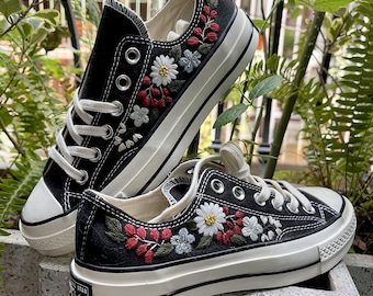 May include: Black canvas Converse sneakers with white soles and embroidered floral designs in red, white, and green.