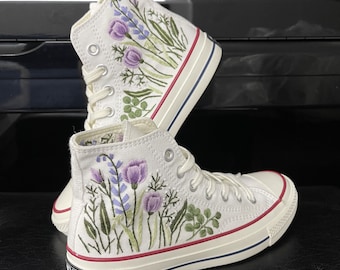Pode incluir: Tênis branco de lona de cano alto com flores bordadas em roxo e verde. Os tênis têm uma sola de borracha branca com uma faixa vermelha.