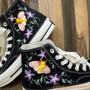 Könnte beinhalten: Paar schwarze High-Top-Sneaker mit weißen Schnürsenkeln und Sohlen. Die Sneaker haben gestickte Designs einer rosa und gelben Motte, lila Blumen und grüne Ranken. Die Schuhe haben einen klassischen, lässigen Stil.