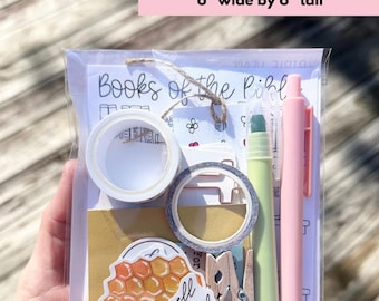 May include: A clear plastic bag containing stationery items. Includes decorative tape, stickers, a green highlighter, a pink pen, and wooden clothespins. The bag is labeled "Bible Grab Bag" and measures 6 inches wide by 6 inches tall.