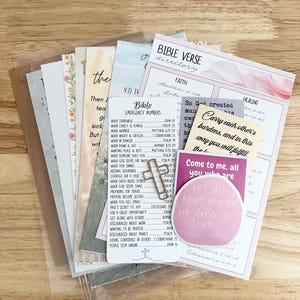 May include: A collection of Bible verse cards and stickers, featuring various designs and inspirational quotes. The cards include a Bible verse directory, emergency numbers, and a metal cross. The color palette includes pink, white, and light blue.
