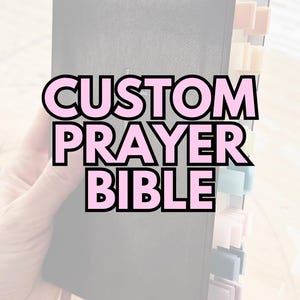 May include: A black Bible with colorful page tabs and a pink ribbon bookmark. The text "CUSTOM PRAYER BIBLE" is displayed in pink letters with a black outline. The Bible is held in a person's hand.
