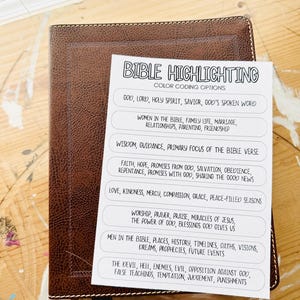 May include: A brown leather Bible with white stitching and a color-coded Bible highlighting guide. The guide lists color-coding options for topics such as God, faith, love, and the devil. The Bible is on a wooden surface.