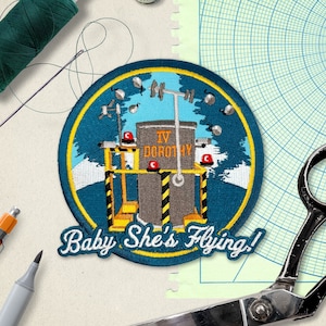 May include: A blue and yellow embroidered patch with a rocket launch scene. The patch has the text "IV DOROTHY" and "Baby She's Flying!"