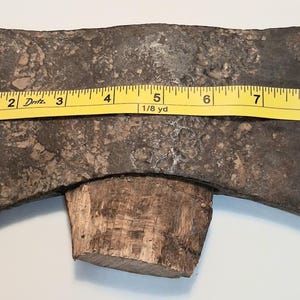 Op de afbeelding: Een dubbelzijdige bijlkop met een donkere, verweerde en gepitte metalen afwerking, voorzien van een kort houten handvat. Een geel Dritz-meetlint is over het blad geplaatst en toont een totale breedte van ongeveer 23,5 centimeter.