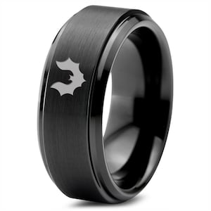 Puede incluir: Un anillo de carburo de tungsteno negro con un borde biselado y un acabado cepillado. El anillo presenta un pequeño diseño de murciélago grabado.