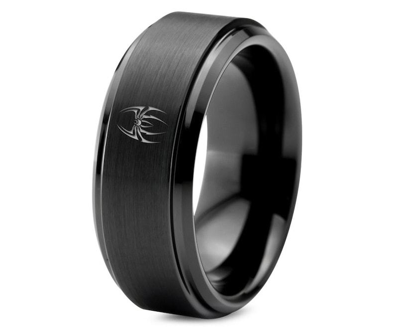 May include: A black tungsten carbide ring with a beveled edge and a brushed finish. The ring features a stylized spider design engraved on the top.