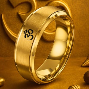 May include: A gold-colored ring with a brushed metal finish. The ring features a black Om symbol engraved on the band. The ring has a polished inner surface and a rounded profile, reflecting light.