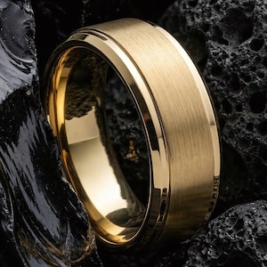 Könnte beinhalten: Ein goldfarbener Ring mit gebürsteter Metalloberfläche. Der Ring hat ein flaches, breites Band mit polierter Innenkante. Der Ring ist vor einem dunklen, strukturierten Hintergrund platziert.