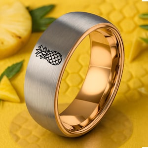 May include: A two-tone ring with a brushed silver exterior and a gold interior. A black pineapple design is etched on the silver band. The ring is set against a yellow background with pineapple slices.