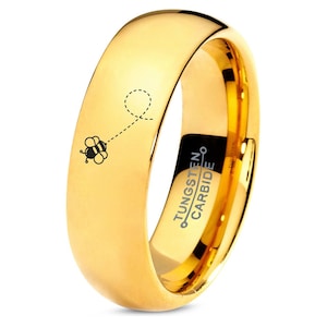 May include: A gold tungsten carbide ring with a bee and dotted line design engraved on the band. The inscription "TUNGSTEN CARBIDE" is engraved on the inside of the ring.