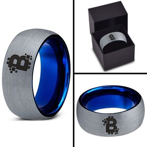 Op de afbeelding: Een zilveren ring met een blauwe binnenband en een zwart Bitcoin-symbool gegraveerd op de bovenkant. De ring zit in een zwarte doos.